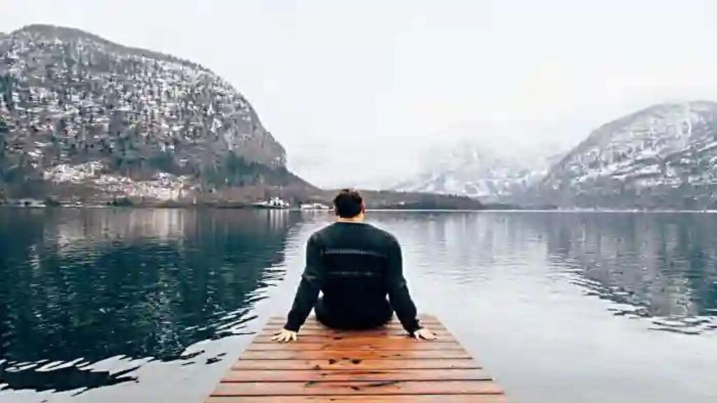 Como ter mais Foco para Alcançar seus Objetivos? A imagem mostra um rapaz, de costas, sentando num deck a beira de um lago, ao fundo duas montanhas com um pouco de neve.