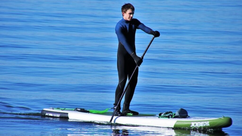 Tire Proveito do seu Estilo de Vida. No mar, nas águas mansas, um rapaz remando em pé na sua prancha stand up paddle. Ele está usando roupa de mergulho, na cor preta com detalhes em azul pavão.
