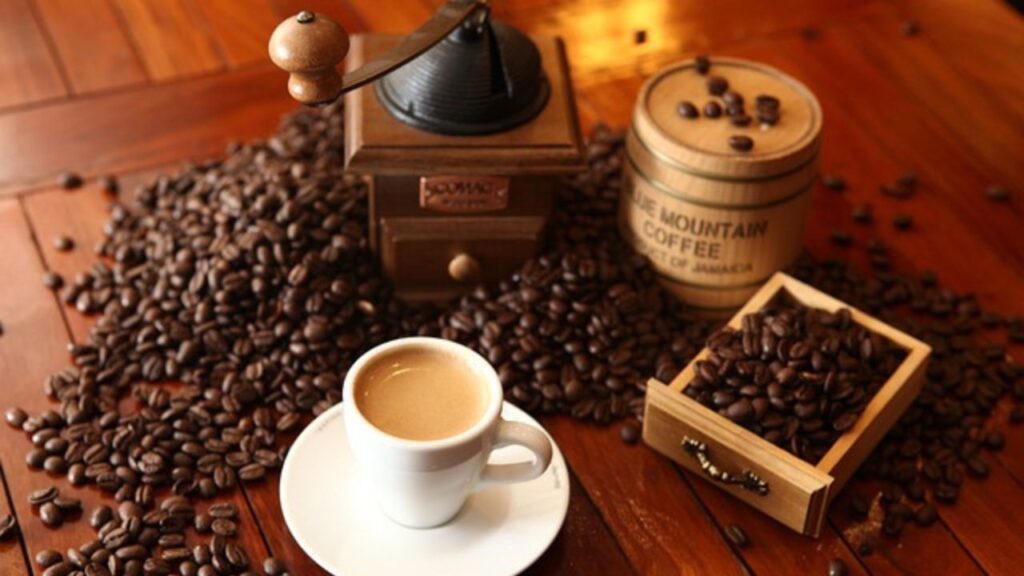 Quais os Benefícios do Café para o Cérebro? Um pires e uma xícara brancos, no seu interior café cremoso e quentinho, sobre uma mesa de madeira ao lado de grãos torrados de café e uma mini trituradora de grãos.