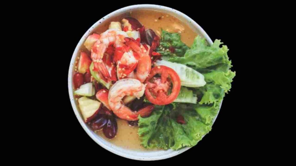 Uma tigela branca com um suculento ensopado rico em nutrientes, como: alface, tomate, pepino, uva rose, frutos-do-mar, pedaços de maçã sem sementes e com a casca, salpicado por gergelim.