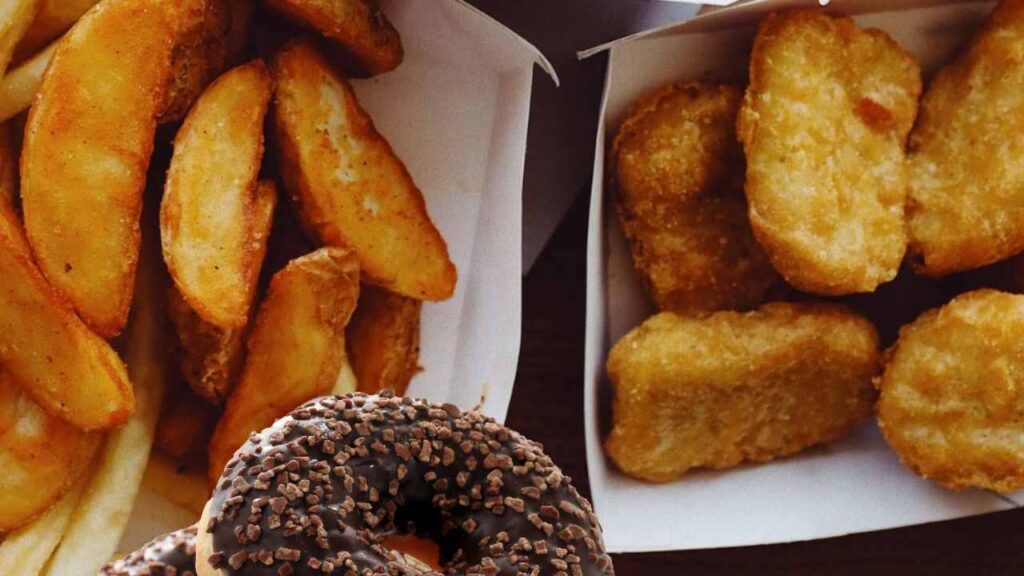 Vários salgadinhos fritos em óleo vegetal, rico em gordura trans, separados em diferentes recipientes de papelão para absorver a gordura e ao lado uma rosca de glúten coberta com achocolatado rico em açúcar.