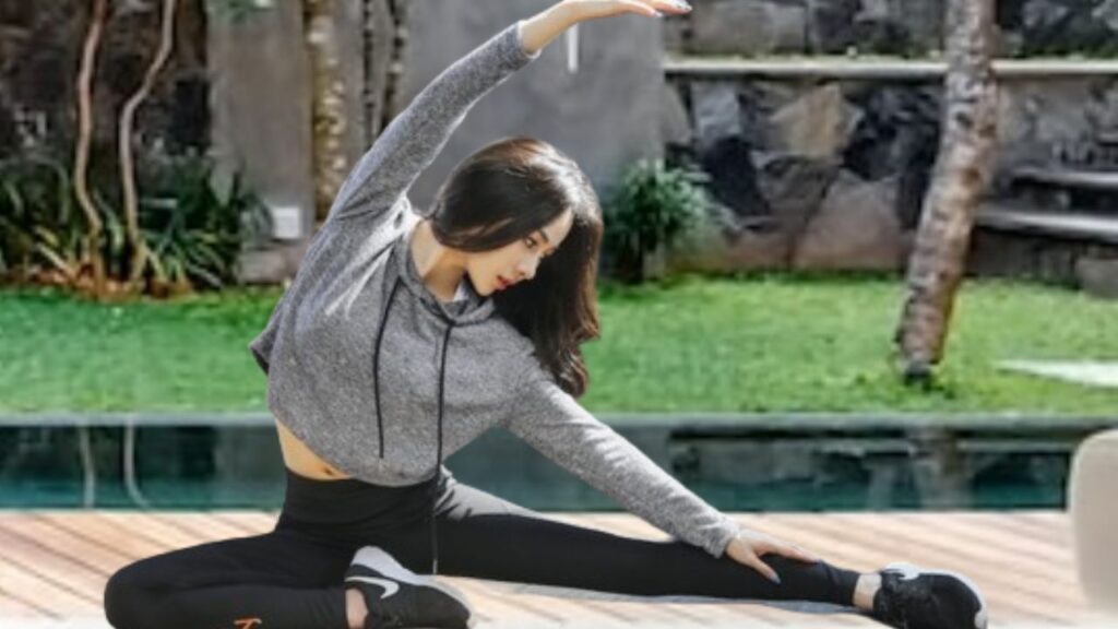 Exercícios em Casa Sem Equipamentos: Guia Completo. Na imagem, uma moça, de origem oriental, pratica Yoga na varanda da sua casa, a beira da piscina. Ela está usando uma calça “legging” na cor preta, usando um tênis preto e uma camiseta com capuz na cor cinza mesclada.